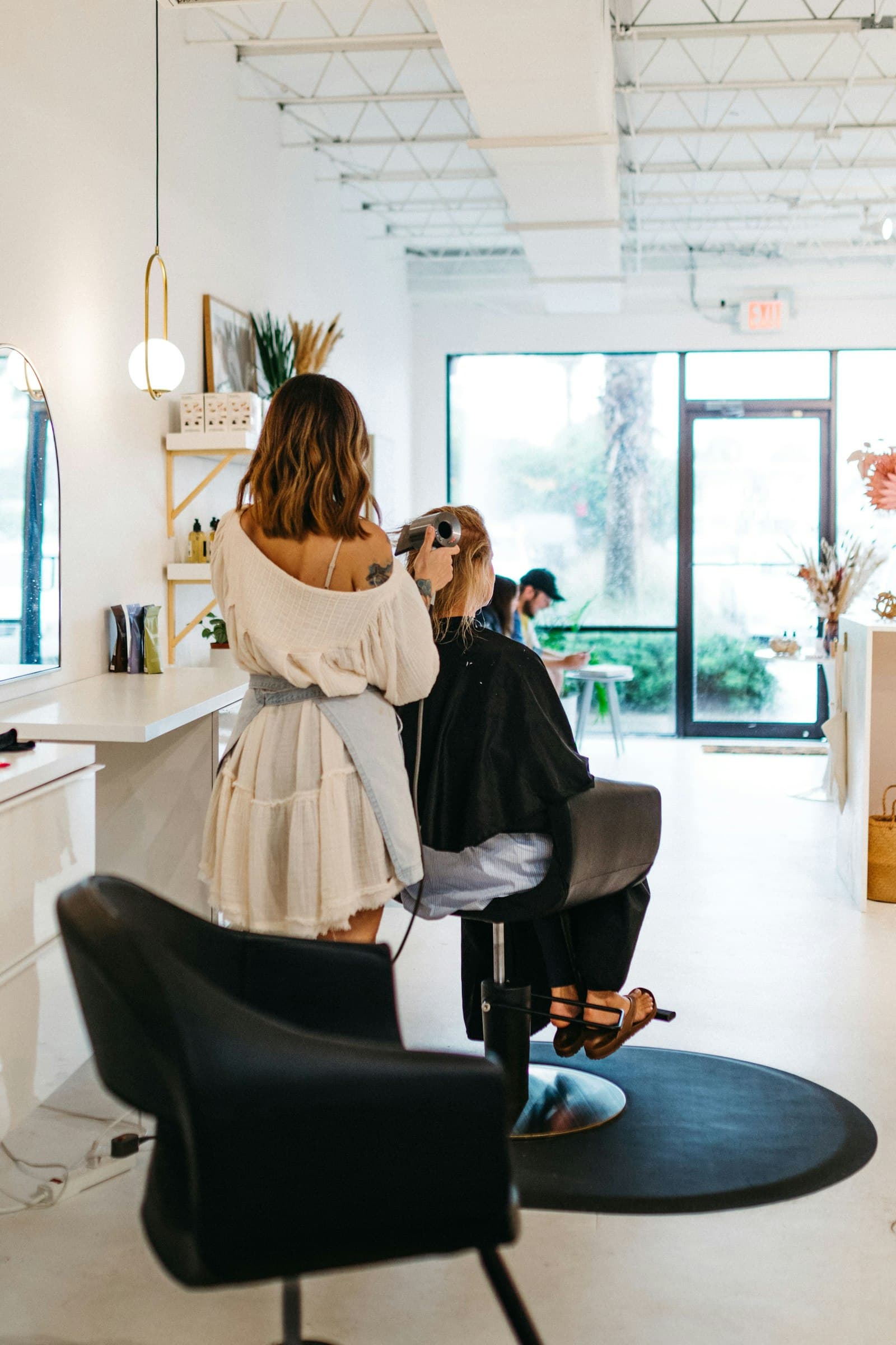 A stylist finishing a client’s hair in a bright salon with styling chairs and a big front window